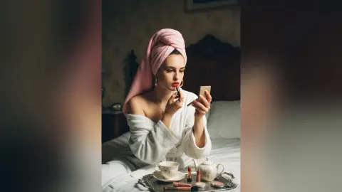 Young woman in white bathrobe and pink hair towel applying lipstick in a cozy bedroom setting.