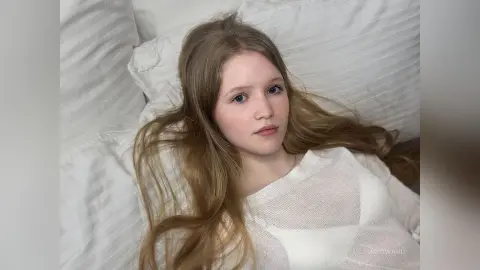 Young woman with long blonde hair wearing a white knit sweater relaxing on white pillows in a bright bedroom.