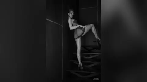Elegant woman in a dark bodysuit posing on stairs in a moody, cinematic black and white setting.