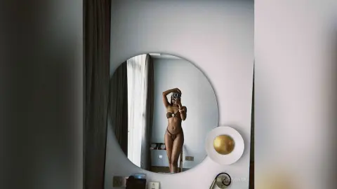 Slim woman in a black bikini taking a mirror selfie in a bright, modern minimalist bedroom.