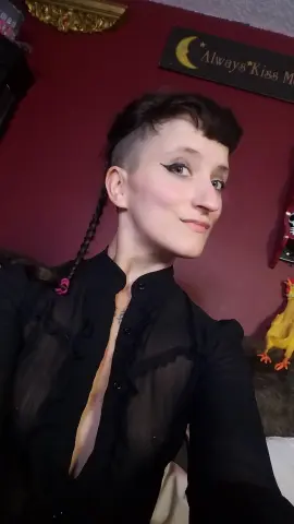 Smiling woman with short dark hair and winged eyeliner wearing a black sheer blouse in a cozy room.