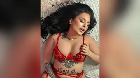Tattooed woman in red lace lingerie relaxing on a white bed with a serene mood.