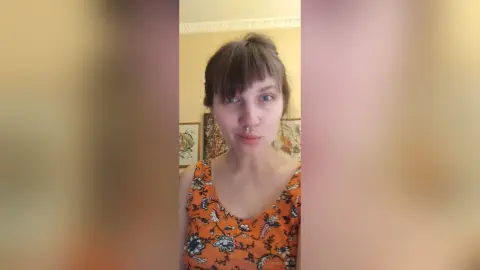 StacyMalvin looks at the camera wearing an orange floral tank top with her brown hair styled in a bun and blunt bangs.