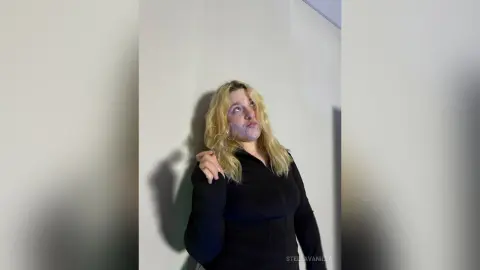 StellaVanilla with long blonde hair wearing a black long-sleeve top, looking up and making a face against a plain white wall.
