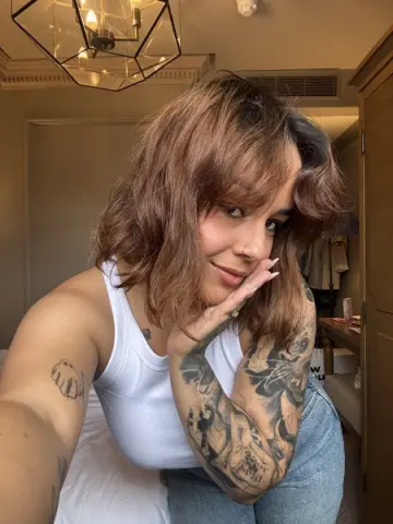 Smiling woman with wavy brown hair and arm tattoos wearing a white tank top in a cozy bedroom setting.