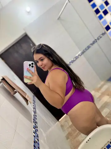 Curvy woman in a purple swimsuit taking a selfie in a modern tiled bathroom.