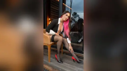 Young woman with pink hair in a black and white dress and red heels sitting on a wooden deck at sunset.