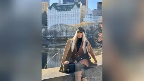 Stylish woman in a brown trench coat and black turtleneck sitting by a city lake on a sunny day.