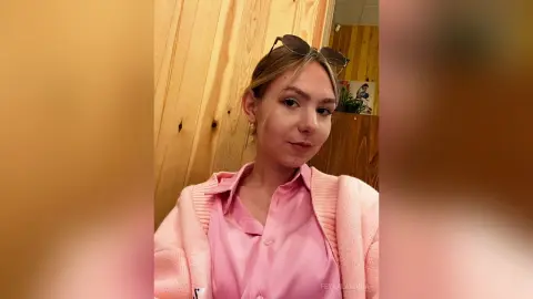 Young woman in a pink button-down shirt and cardigan posing in a warm wooden interior.