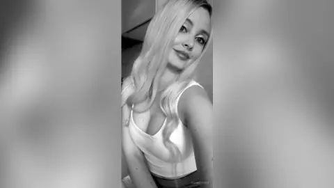 DianeDavies poses in a white tank top, showing off her long blonde hair and cleavage in this black and white photo.