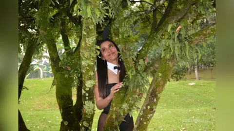 AmeliaStell poses behind tree trunks in a park wearing a black and white maid outfit.