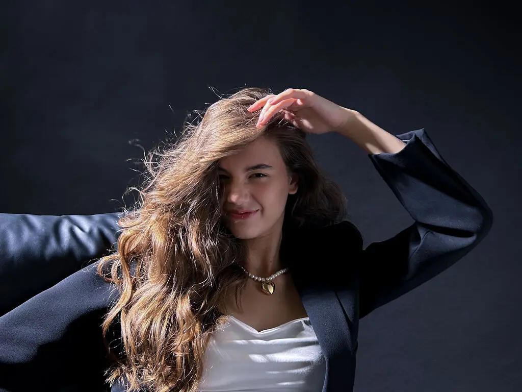 Smiling woman with long wavy hair wearing a black blazer and pearl necklace against a dark studio background.