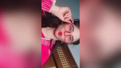 Young woman in a pink lace top wearing glasses with a playful expression in a cozy indoor setting.