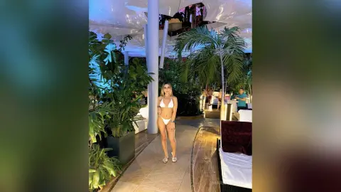Blonde woman in a white bikini standing in a lush tropical indoor garden setting with a relaxed summer mood.