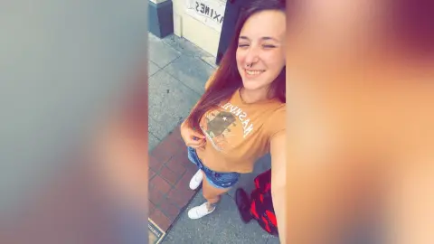 Smiling young woman in a mustard graphic t-shirt and denim shorts standing on a city sidewalk.