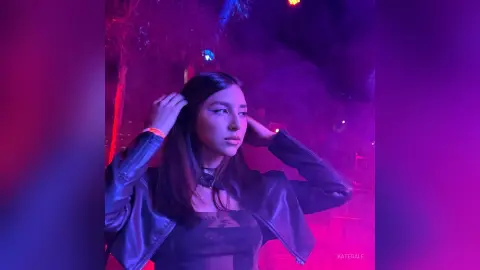 KateGale poses with her hands in her long dark hair, wearing a black leather jacket and black top against a purple-lit background.