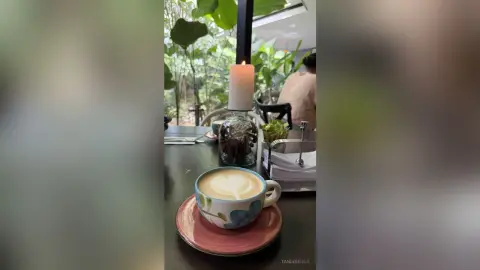 Latte art in a ceramic cup on a cafe table with a lit candle and lush green plants in a cozy setting.