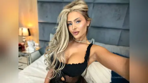 Glamorous blonde woman in a black ruffled top posing in a cozy bedroom setting.