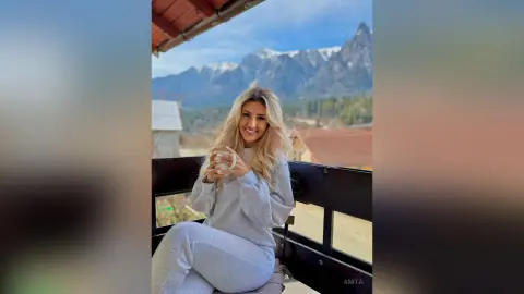 Anita smiles while sitting on a balcony overlooking snow-capped mountains, wearing a light grey long-sleeve lounge set.