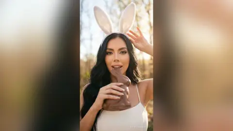 Woman in white bunny ears eating a chocolate bunny in a sunny outdoor setting with a playful mood.