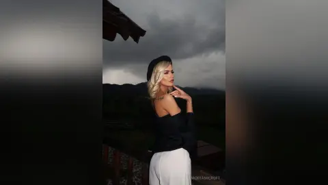 Blonde woman in a black top and white trousers posing against a moody, cloudy mountain landscape.