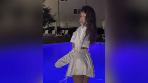 Young woman in a white satin outfit posing by a glowing blue swimming pool at night.