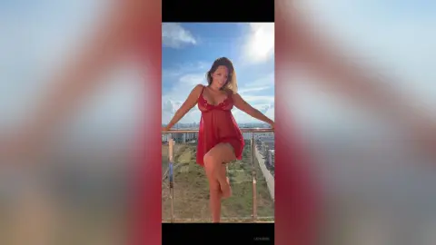 Smiling woman in a red lace chemise posing on a sunny balcony overlooking a city skyline.
