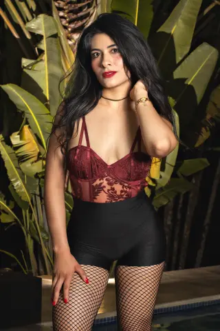 Young woman in burgundy lace corset and black shorts posing amidst lush tropical green leaves.