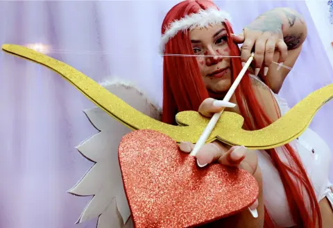 Cosplayer in white angel outfit with red hair holding a glittery heart and gold wings in a dreamy setting.