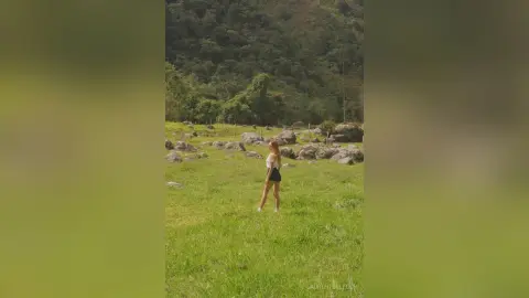 LaurentBelluci stands in a grassy field wearing a white top and black shorts against a backdrop of lush green hills.