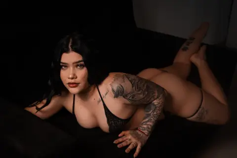 Madisson_Lee20 lies on a black couch wearing black lace lingerie, showing off her tattoos and dark hair.
