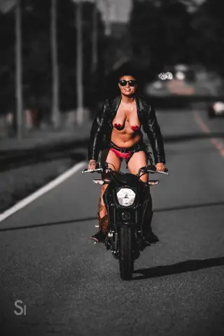 Confident woman in a black leather jacket and red bikini riding a black motorcycle on a sunny city street.