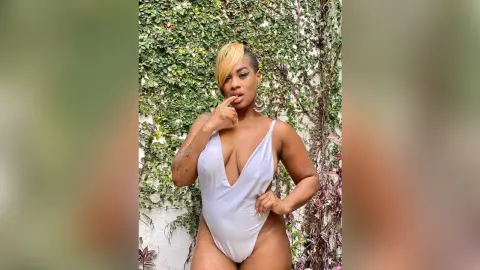 Confident woman in a white plunging bodysuit posing against a lush green ivy wall.