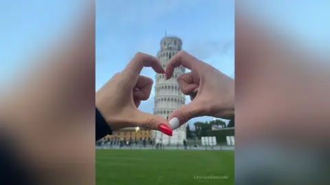 Hands forming a heart shape in front of the Leaning Tower of Pisa under a soft blue sky.