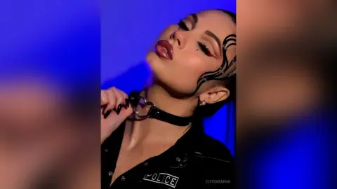 CutemEmma poses with a black choker and black police shirt, looking up with red eyeliner and dark hair pulled back against a blue background.