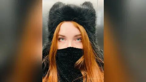 Redheaded woman wearing a cozy black faux fur hat and face mask in a modern setting.