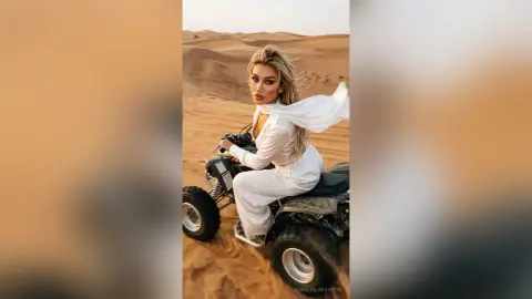 Stylish woman in white jumpsuit riding an ATV through golden desert sand dunes at sunset.