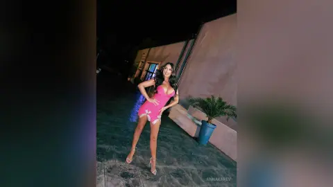 AnnaKarev poses in a tight pink mini dress and high heels outside at night.