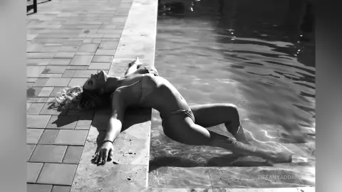TiffanyAddams lies on the edge of a pool in a light bikini, arching her back with her head and arms resting on the stone tiling.