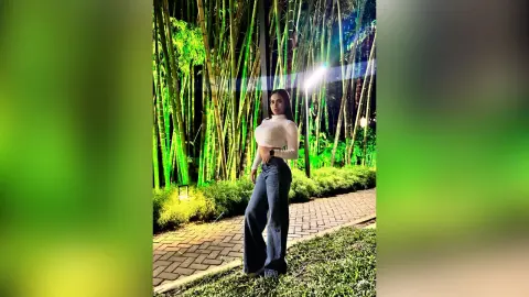 MiaKorz stands outdoors at night in front of tall bamboo stalks, wearing a white long-sleeved crop top and dark blue flared jeans.