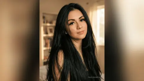 Young woman with long black hair in a warm, sunlit indoor setting with a serene expression.