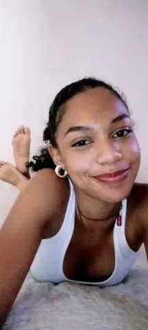 Smiling young woman in a white tank top relaxing on a bed in a bright, minimalist room.