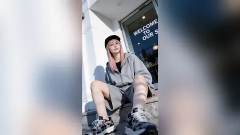 Young woman in grey hoodie and black cap sitting outdoors by a building entrance in bright sunlight.