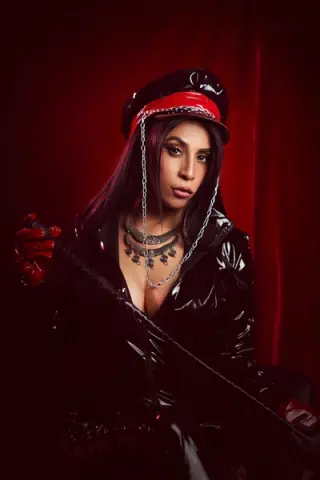 Glamorous woman in black latex outfit and captain hat posing against a moody red velvet backdrop.