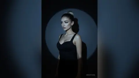 Elegant woman in a black tank top posing under dramatic spotlight in a moody, dark studio setting.
