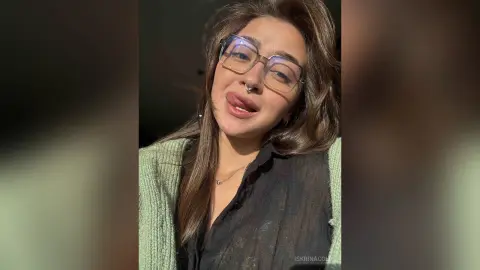 Young woman with glasses and nose ring wearing a black shirt and green cardigan in soft natural lighting.