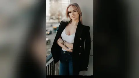 Smiling blonde woman in a black blazer and white tank top posing on a city balcony during golden hour.