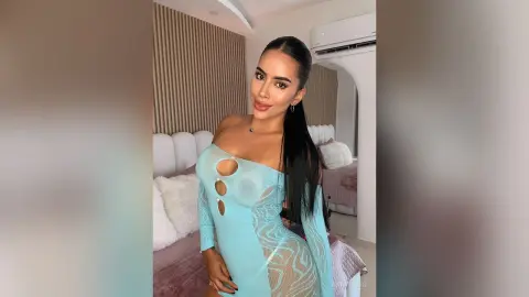 Glamorous woman in a light blue cutout bodycon dress posing in a modern, bright bedroom.