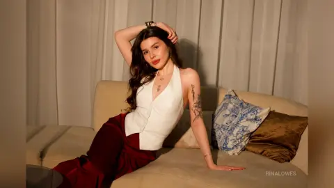Stylish woman in a white sleeveless top and red trousers relaxing on a beige sofa in a warm, cozy setting.