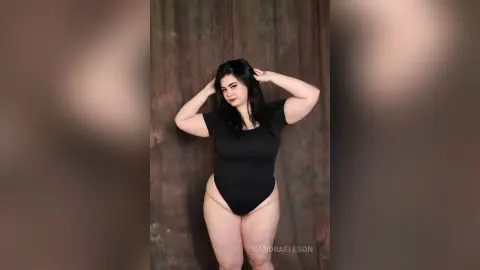 Confident woman in a black bodysuit posing against a rustic textured backdrop.